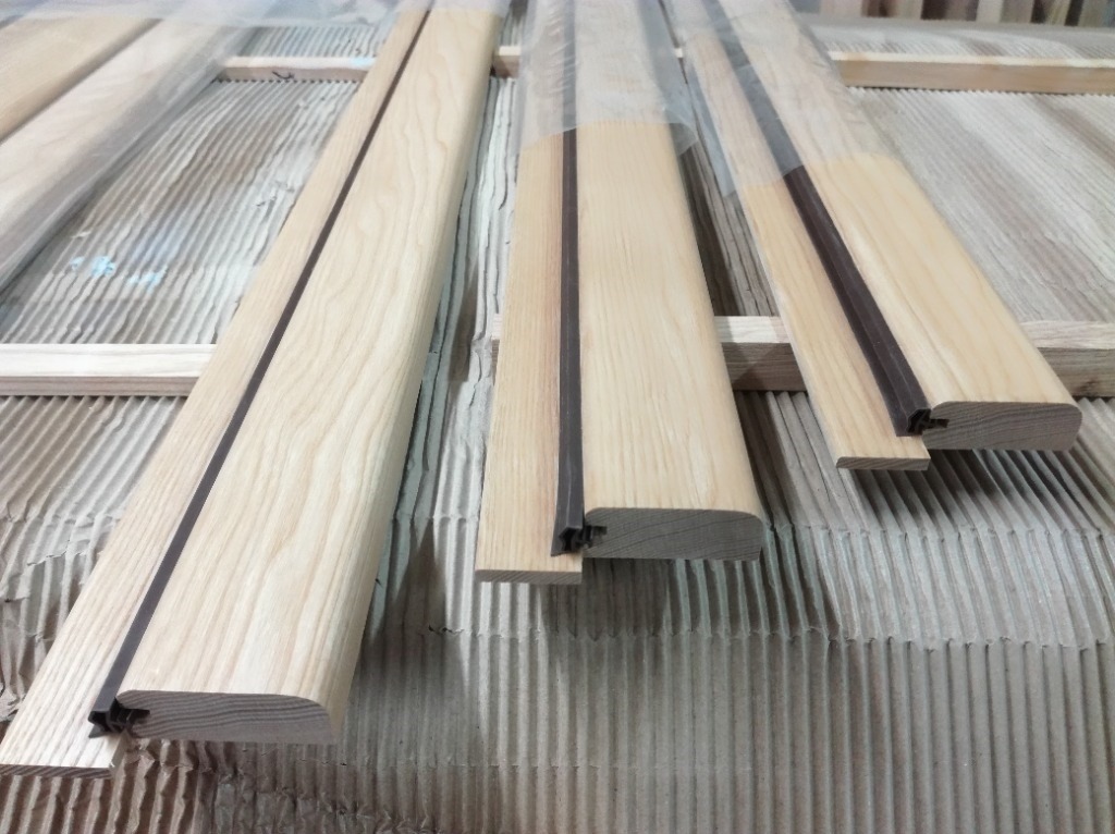 Elementy drewniane, prawdopodobnie ościeżnice lub ramiaki, z widocznym profilem i uszczelką, ułożone na tekturze falistej.