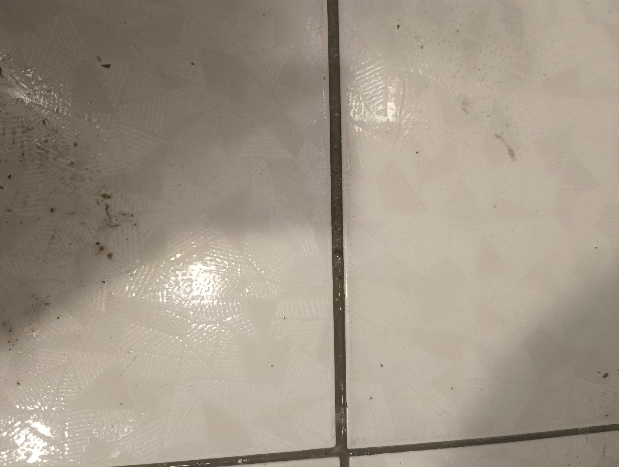 Białe płytki podłogowe z wyraźnymi śladami brudu i plam przed czyszczeniem, widoczne fugi.