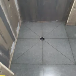 Szare płytki podłogowe ułożone w kwadraty zbiegające się do odpływu liniowego w kabinie prysznicowej, ściany przygotowane pod okładzinę, widoczne żółte elementy instalacji hydraulicznej.