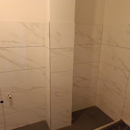 Budownictwo - Ściany łazienki wyłożone białymi płytkami imitującymi marmur, widoczne rury instalacji wodnej przed montażem armatury.