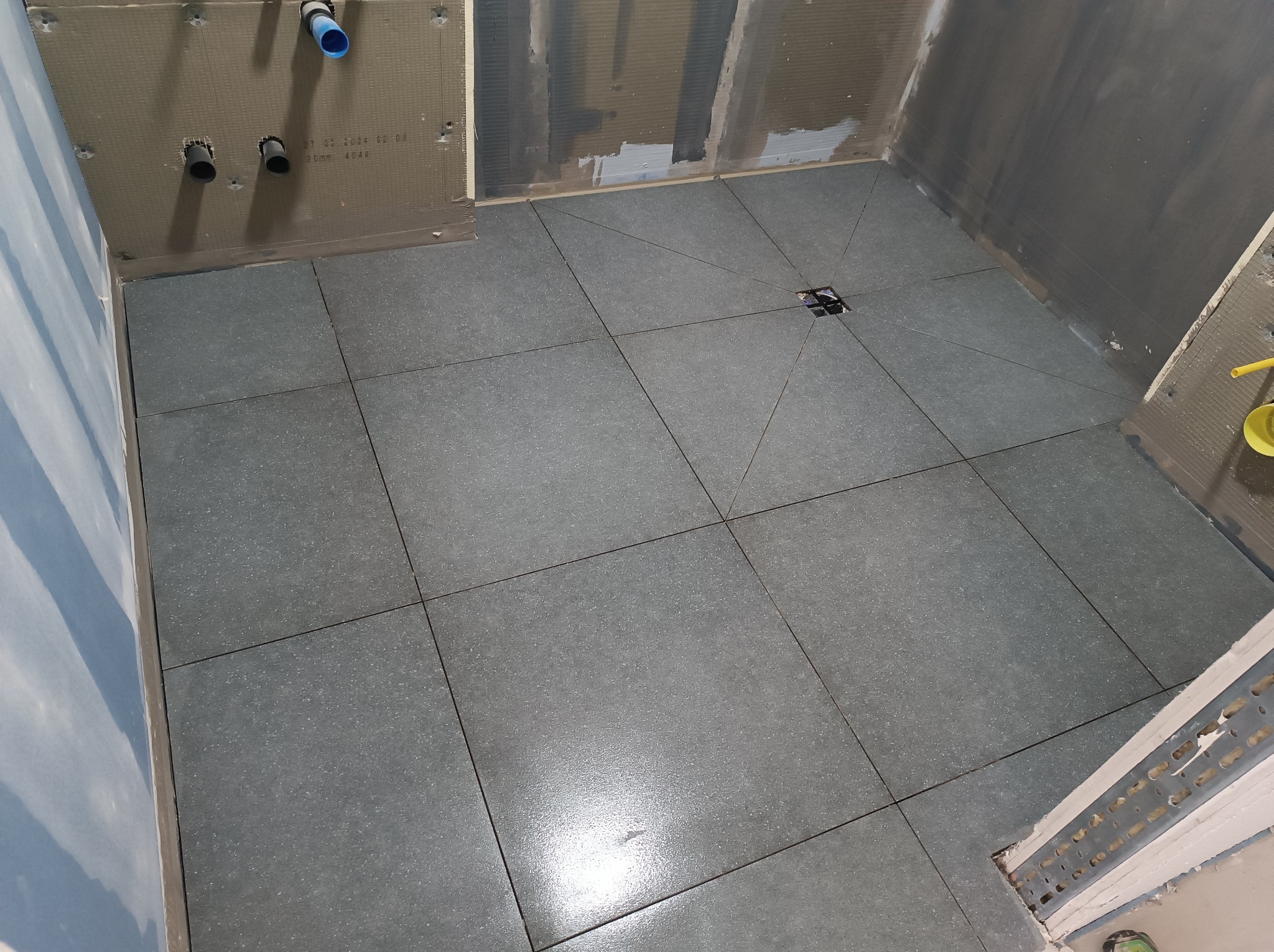 Ułożone, ciemnoszare płytki podłogowe w łazience w trakcie remontu w Świerzawie. Widoczny odpływ liniowy, rury instalacji wodnej oraz niezabezpieczone ściany przygotowane do dalszych prac...