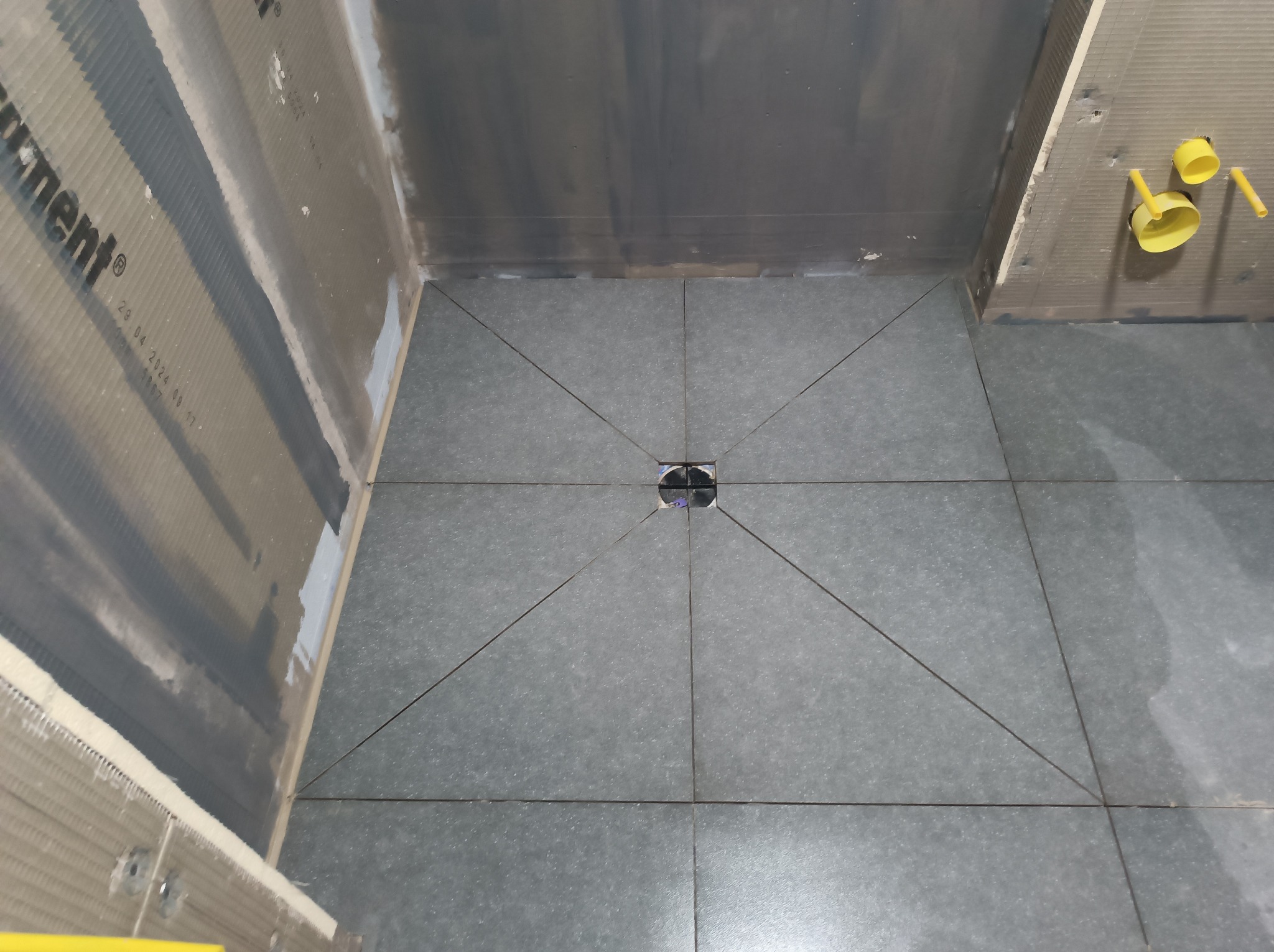 Szare płytki podłogowe ułożone w kwadraty zbiegające się do odpływu liniowego w kabinie prysznicowej, ściany przygotowane pod okładzinę, widoczne żółte elementy instalacji hydraulicznej.