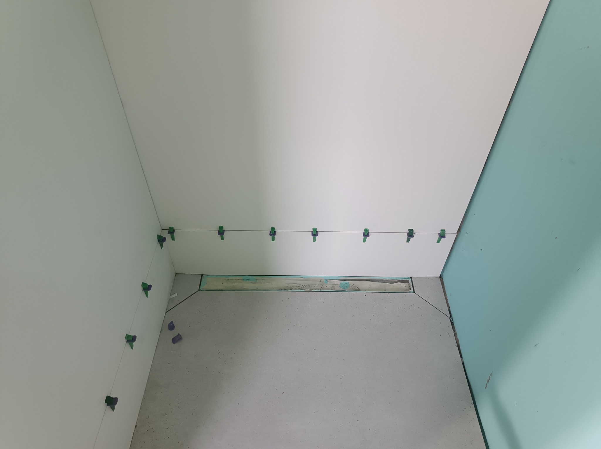 Świeżo położone, białe płytki ścienne w łazience, wyrównane za pomocą zielonych klipsów i sznurka, widoczny odpływ liniowy w szarej posadzce, jedna ściana pomalowana na jasny błękit.