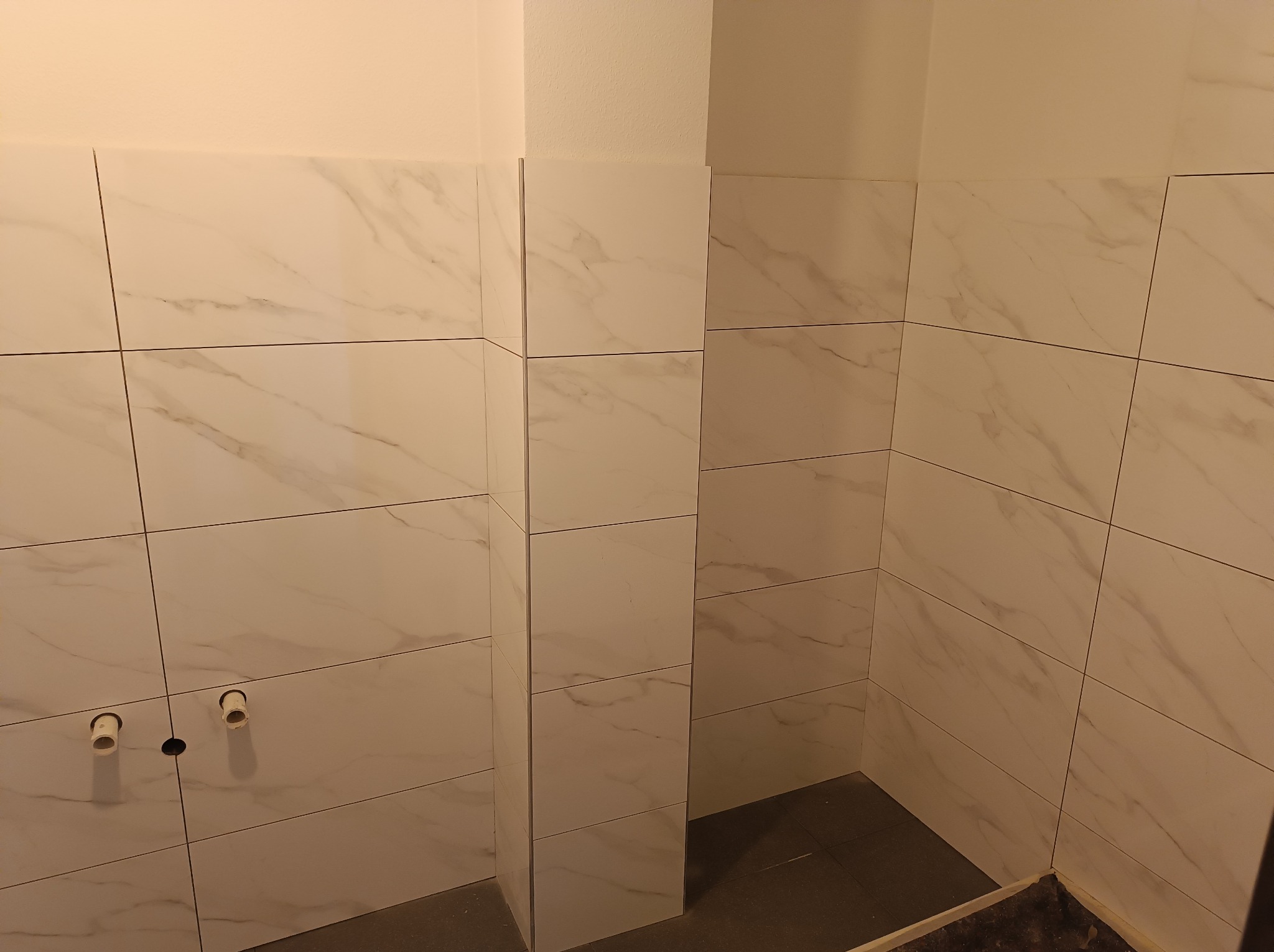 Ściany łazienki wyłożone białymi płytkami imitującymi marmur, widoczne rury instalacji wodnej przed montażem armatury.