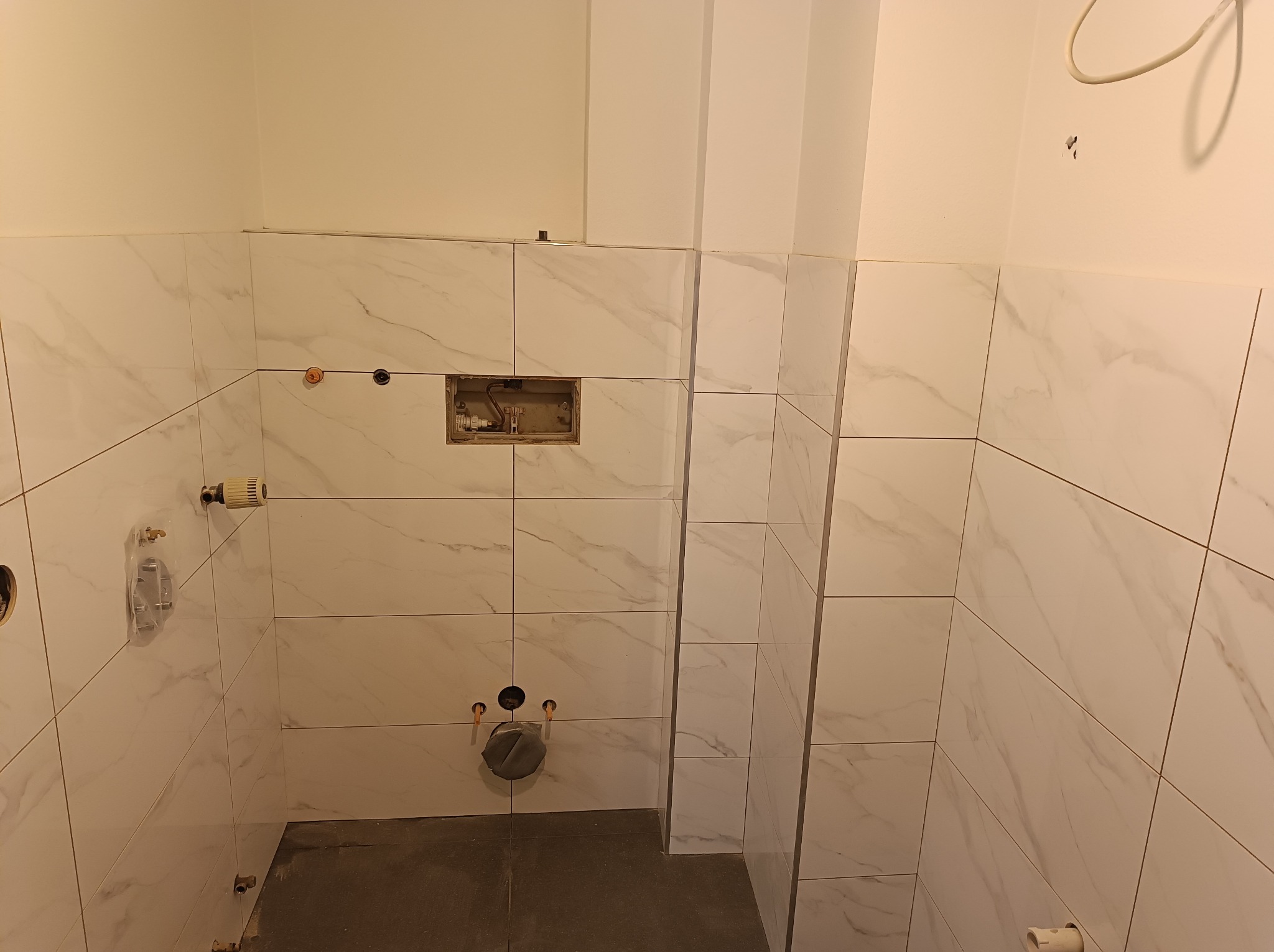 Wnętrze łazienki w trakcie kładzenia białych płytek imitujących marmur, widoczne otwory na rury i instalację podtynkową WC.