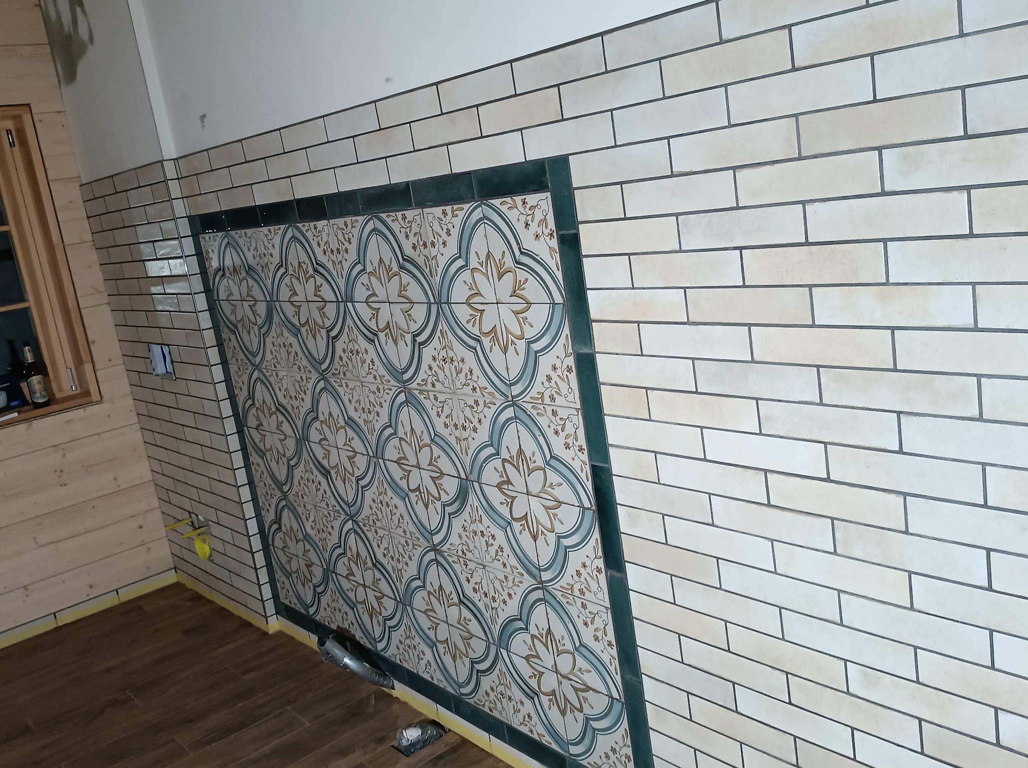 Fragment ściany z ułożoną glazurą w beżowe cegiełki, z dekoracyjnym panelem z płytek w kwiatowy wzór, obramowanym ciemnozieloną glazurą. Podłoga wykończona płytkami imitującymi drewno.