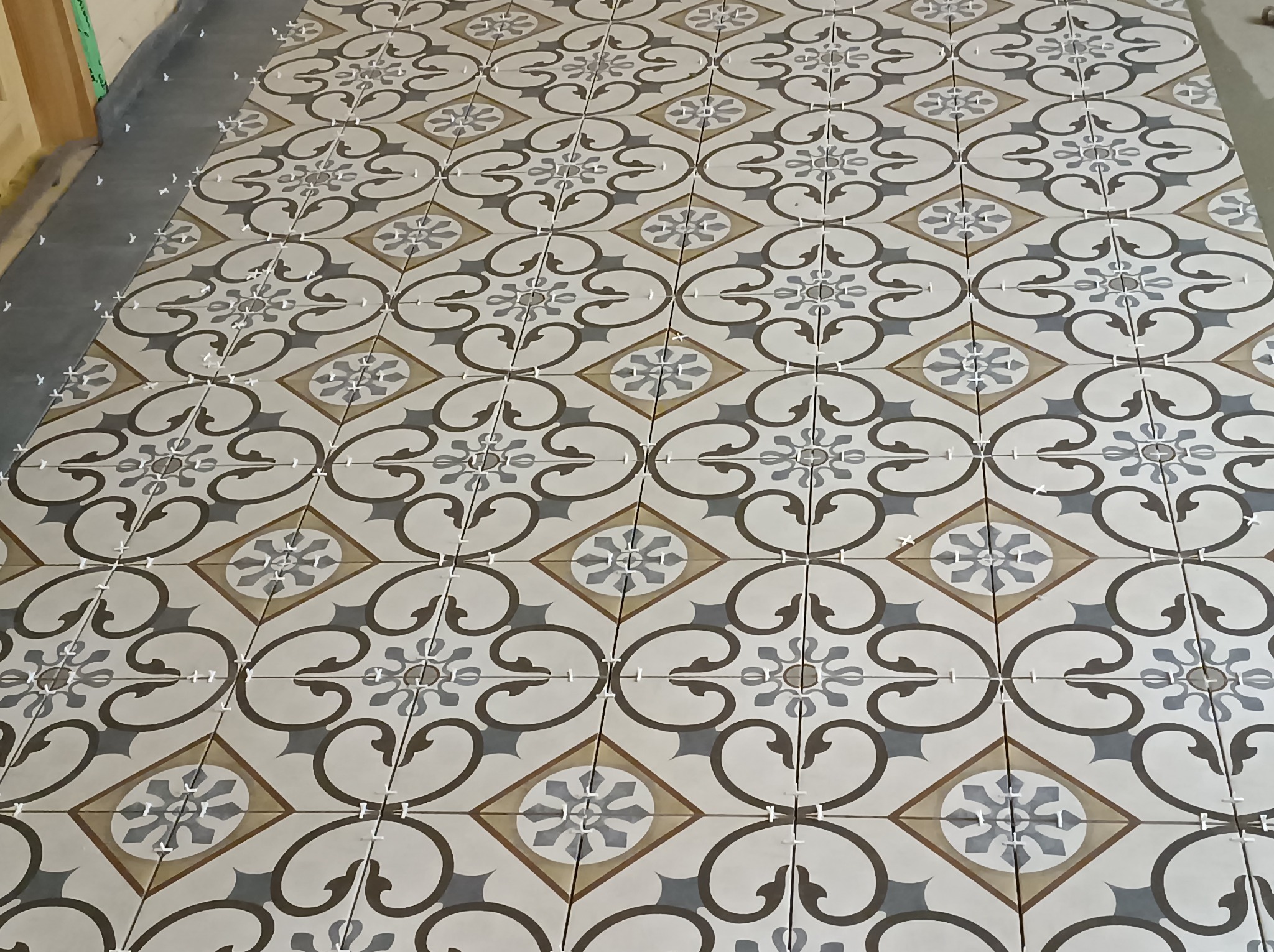 Układanie wzorzystych płytek podłogowych w odcieniach szarości, beżu i bieli z użyciem krzyżyków dystansowych, widoczny fragment podłoża przygotowanego pod fugowanie.