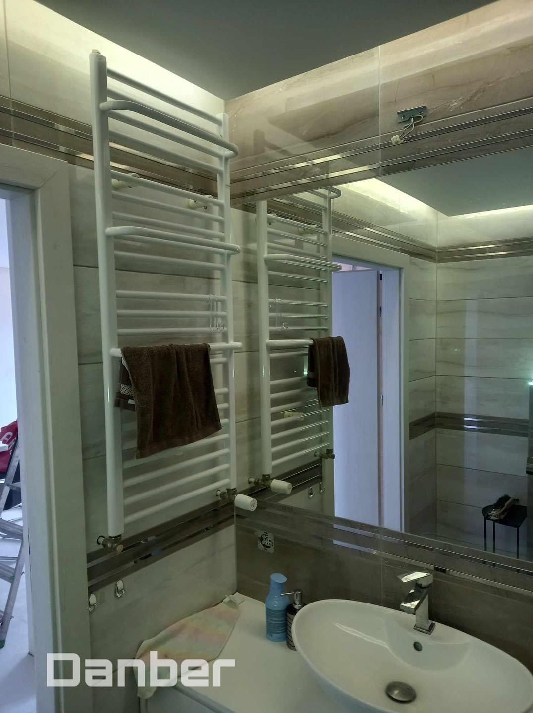 Biały, drabinkowy grzejnik łazienkowy z brązowym ręcznikiem, zamontowany na ścianie z beżowych płytek, widoczny w odbiciu lustrzanym nad umywalką z baterią.