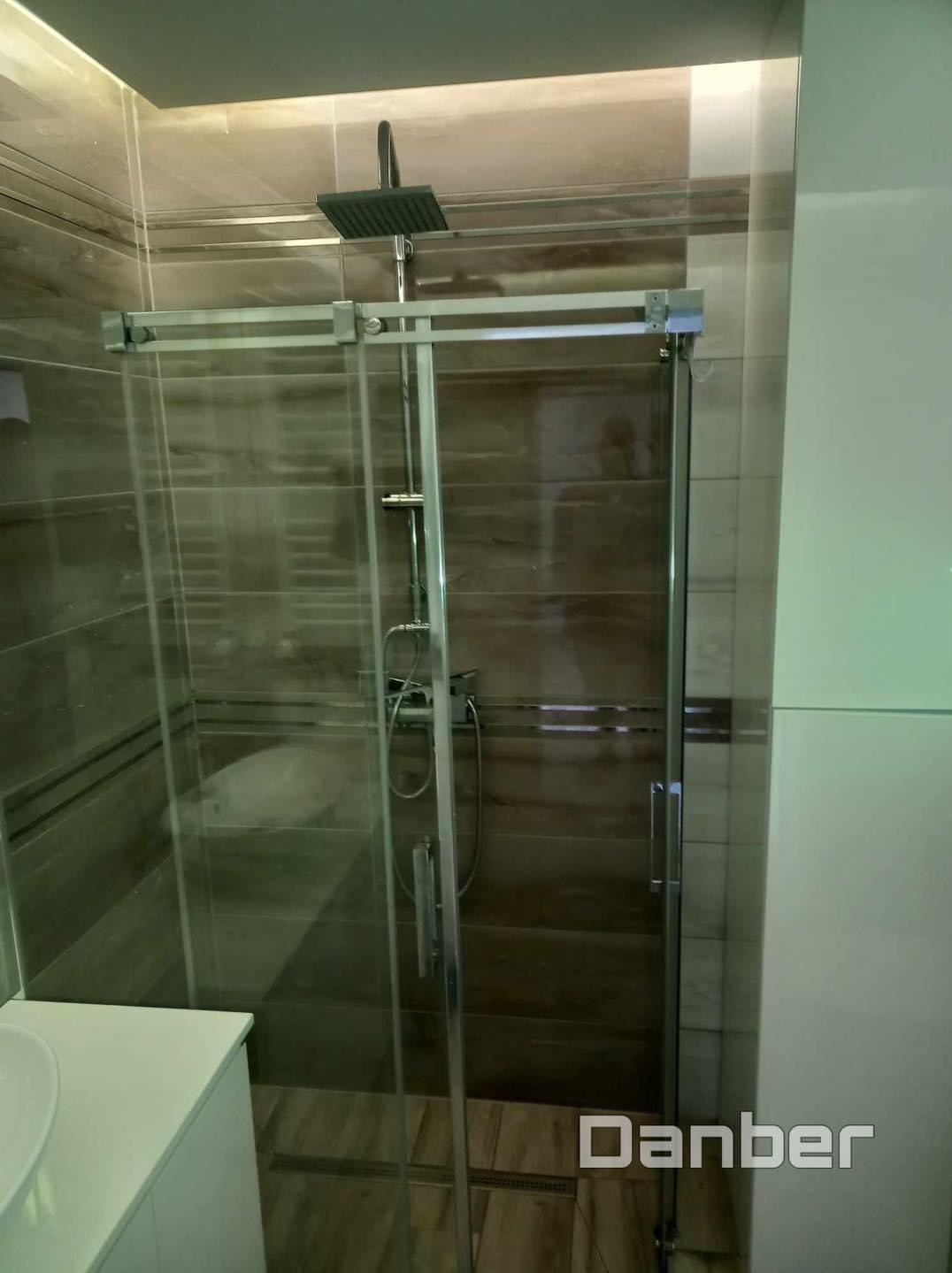 Kabina prysznicowa z przesuwnymi drzwiami i deszczownicą, wykończona brązowymi płytkami imitującymi marmur, z widocznym logo 'Danber' na dolnej części szyby.