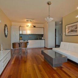 J&D Sp. z o.o. - Jasne wnętrze mieszkania z drewnianą podłogą, białymi meblami i telewizorem z widokiem zachodzącego słońca na ścianie.