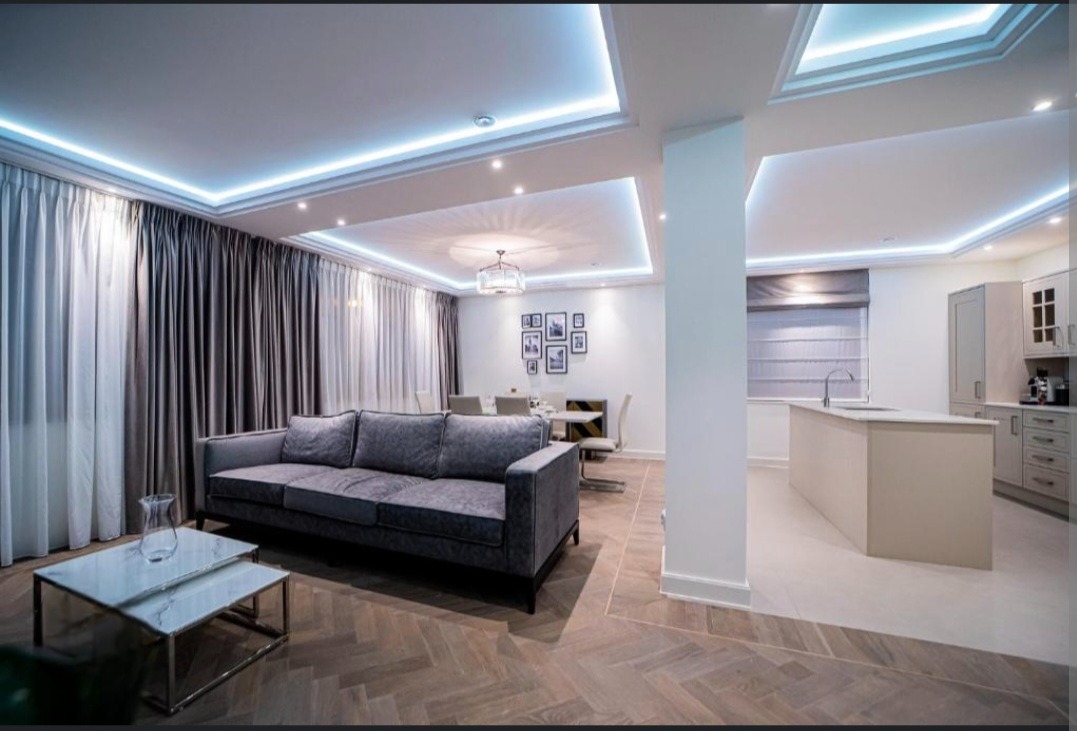 Nowoczesne, przestronne wnętrze mieszkania z szarym narożnikiem, marmurowym stolikiem kawowym, parkietem w jodełkę i oświetleniem LED pod sufitem.