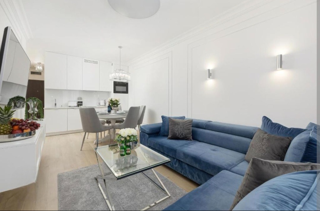 Jasne wnętrze mieszkania z aneksem kuchennym, niebieską sofą narożną i szklanym stolikiem kawowym, z owocami na komodzie w tle.