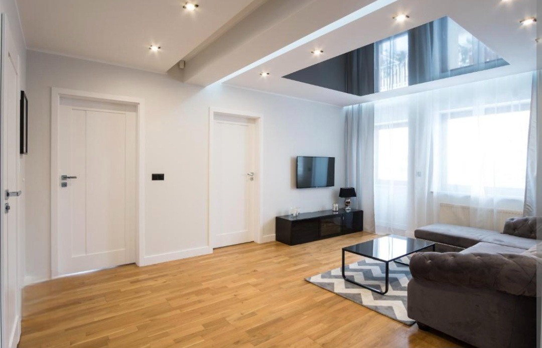 Jasne wnętrze salonu z drewnianą podłogą, szarą sofą narożną, czarnym stolikiem kawowym na wzorzystym dywanie, białymi ścianami i drzwiami, oraz sufitem z oknem dachowym.