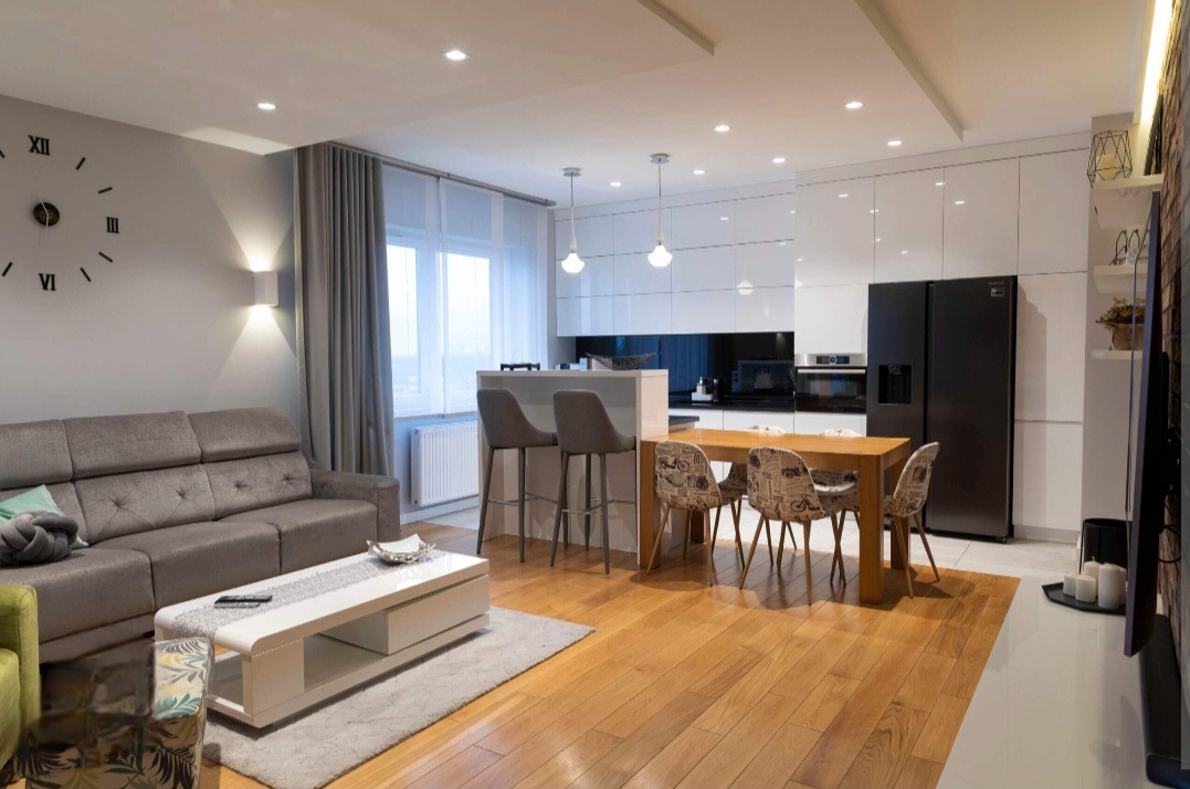 Jasne, przestronne wnętrze mieszkania z drewnianą podłogą, szarą kanapą i białymi meblami kuchennymi, podkreślone przez liczne punkty świetlne.