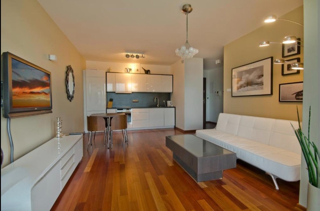 Jasne wnętrze mieszkania z drewnianą podłogą, białymi meblami i telewizorem z widokiem zachodzącego słońca na ścianie.