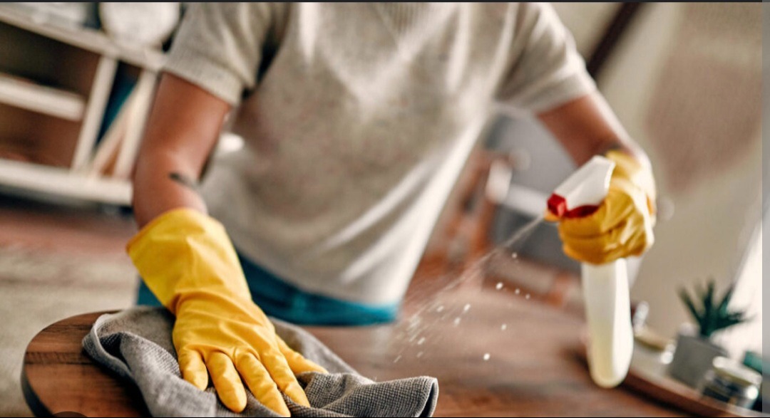 Osoba w żółtych rękawicach spryskuje i czyści drewniany stolik szarą ściereczką, widoczne krople płynu czyszczącego w powietrzu, w tle półka i roślina doniczkowa.