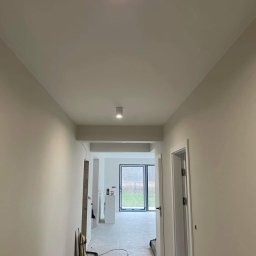 Mykhailo Lavryshyn V&M Construction - Korytarz w trakcie remontu z widocznymi dwoma okrągłymi lampami sufitowymi, świeżo pomalowanymi ścianami w odcieniach bieli, otwartymi drzwiami na końcu korytarza i narzędziami malarskimi opartymi...