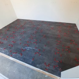 Mykhailo Lavryshyn V&M Construction - Ułożone, ciemnoszare płytki podłogowe imitujące drewno, z czerwonymi klipsami poziomującymi i sznurkami dystansowymi, w trakcie prac wykończeniowych.