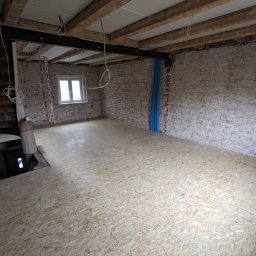 Mykhailo Lavryshyn V&M Construction - Surowe wnętrze w trakcie remontu: płyta OSB na podłodze, odsłonięta cegła na ścianach, widoczne instalacje i drewniane belki stropowe. Okno w tle.