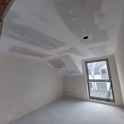 Mykhailo Lavryshyn V&M Construction - Pokój na poddaszu w trakcie szpachlowania: ściany przygotowane, widoczne łączenia płyt gipsowych. Okno z widokiem na dom. Częściowo wykończony sufit.