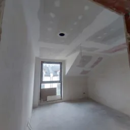 Pokój w trakcie szpachlowania: ściany i sufit pokryte gładzią, widoczne płyty karton-gips, okno i fragment surowego muru przy wejściu. W tle zabudowa jednorodzinna.