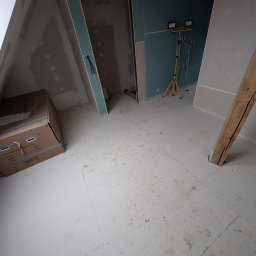 Mykhailo Lavryshyn V&M Construction - Poddasze w trakcie prac wykończeniowych: płyty gipsowe na ścianach, widoczna konstrukcja drewniana, karton na podłodze. W tle lampa budowlana na statywie.
