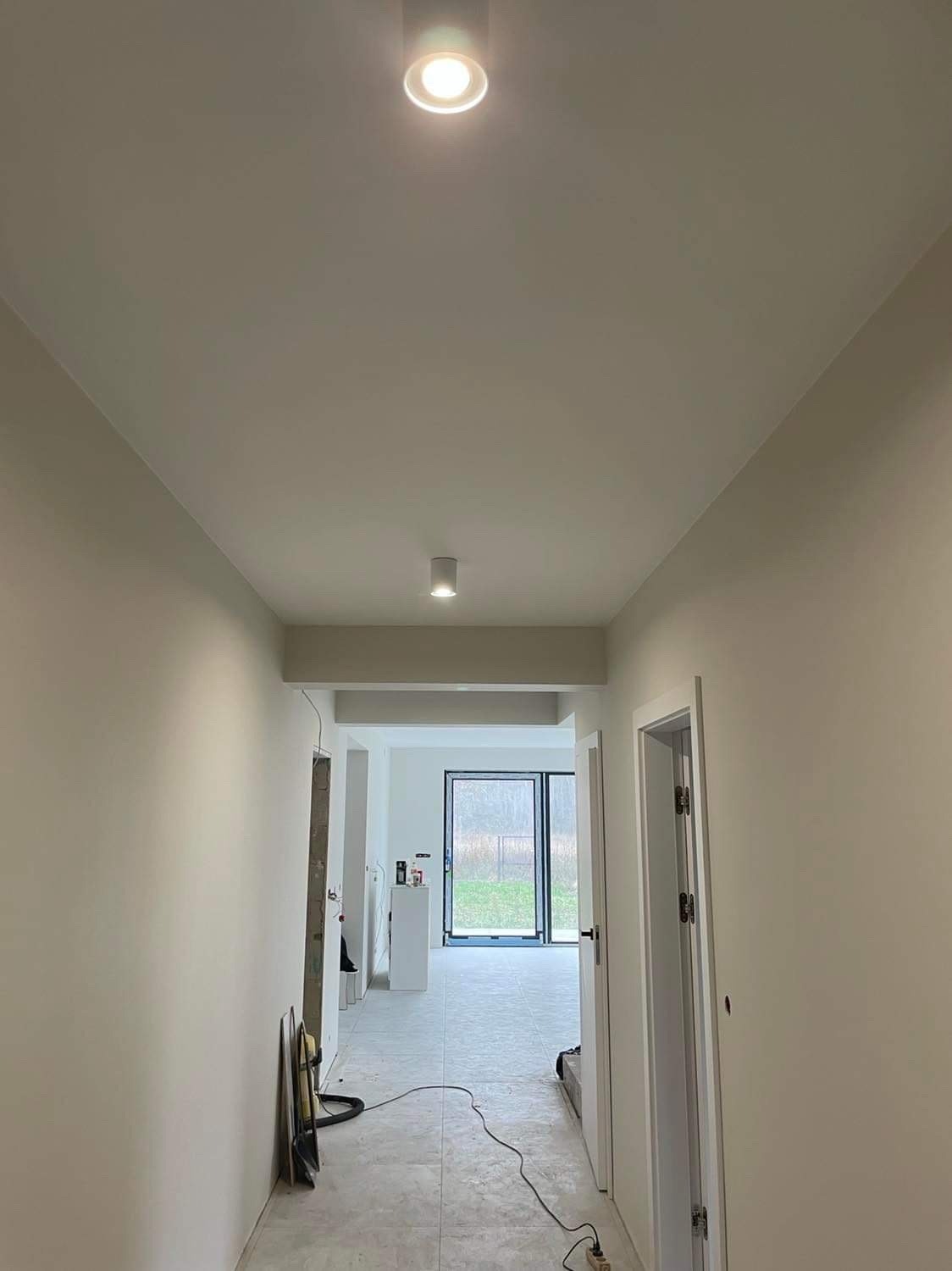 Korytarz w trakcie remontu z widocznymi dwoma okrągłymi lampami sufitowymi, świeżo pomalowanymi ścianami w odcieniach bieli, otwartymi drzwiami na końcu korytarza i narzędziami malarskimi opartymi...