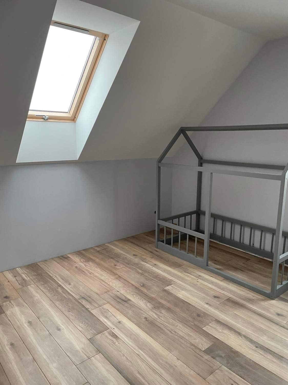 Pokój na poddaszu w Chorzowie po remoncie, z drewnianą podłogą i szarą ramą łóżka w kształcie domku oraz oknem dachowym.