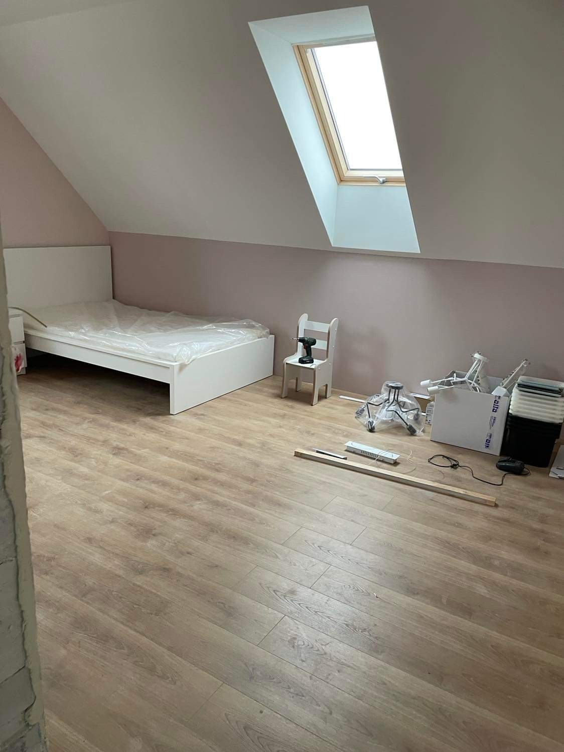 Pokój na poddaszu w trakcie urządzania: łóżko z materacem w folii, drewniana podłoga, okno dachowe, ściany pomalowane na jasny róż, narzędzia i akcesoria montażowe rozłożone na podłodze, wiertarka...