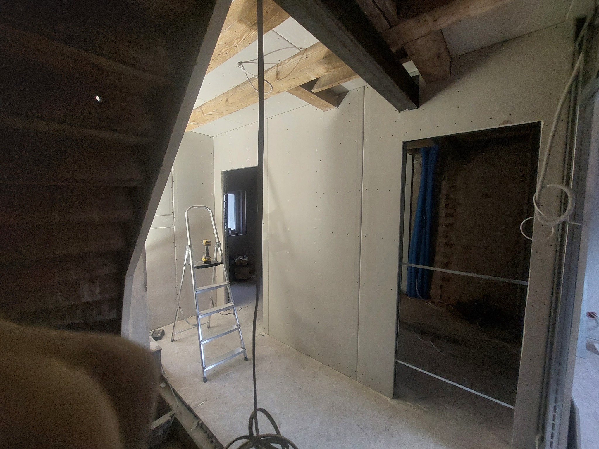 Poddasze w trakcie prac wykończeniowych: zamontowane płyty karton-gips, drewniana konstrukcja dachu, drabina z wkrętarką, widoczne instalacje. Surowy stan, przygotowanie do dalszych prac.