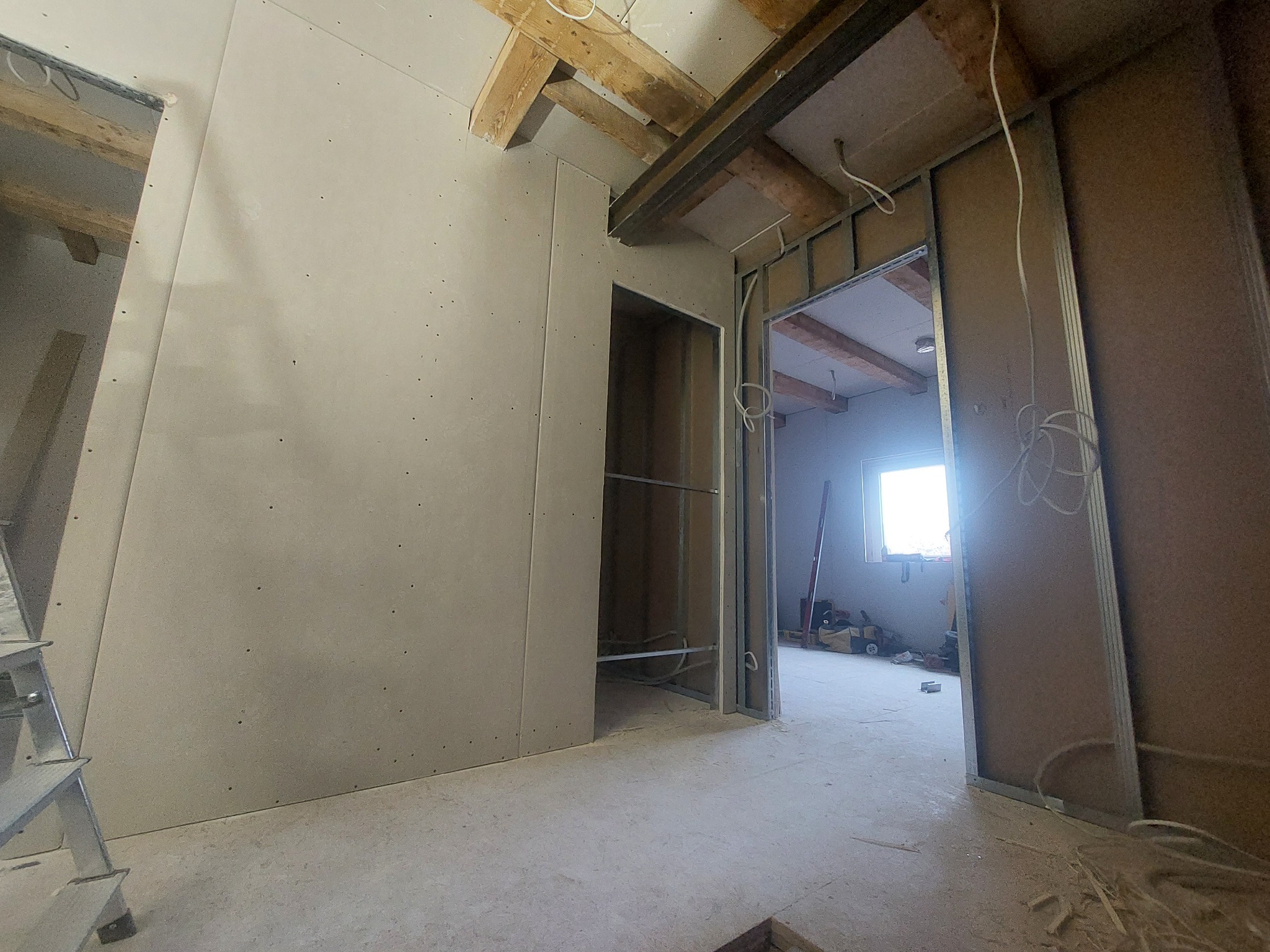 Montaż płyt gipsowo-kartonowych na ścianach i suficie w surowym wnętrzu z drewnianymi belkami. Widoczne metalowe profile i instalacje elektryczne.