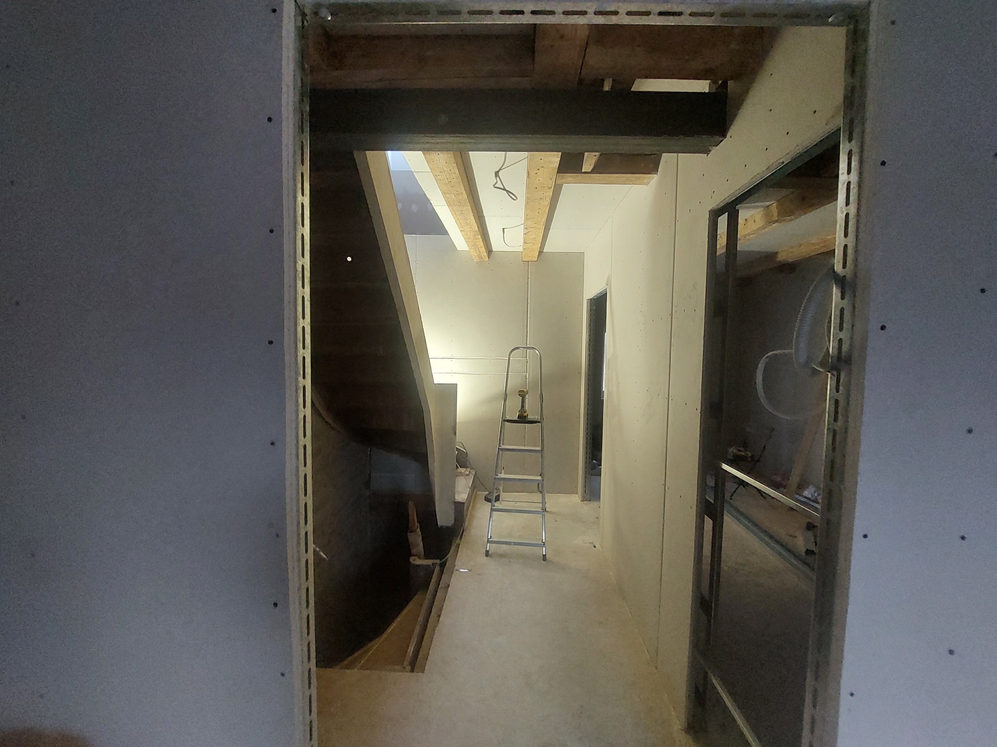 Wnętrze w trakcie wykańczania: surowe ściany z płyt gipsowych, widoczne profile, drabina z wkrętarką w korytarzu, fragment schodów i stropu. Etap prac wykończeniowych.