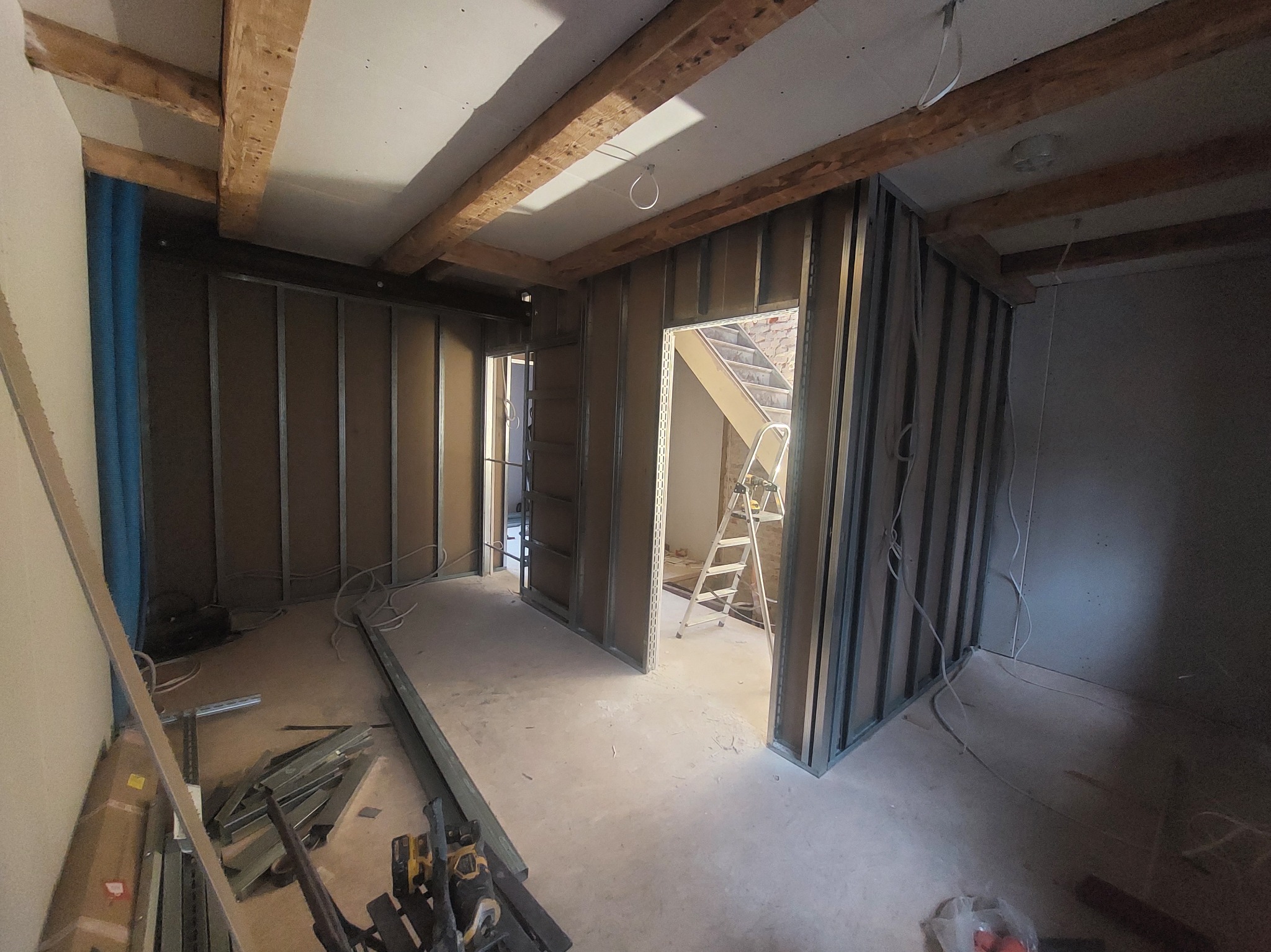 Wnętrze w trakcie montażu ścian działowych z płyt karton-gips, widoczne metalowe profile, drewniane belki stropowe i drabina w przejściu. Etap prac wykończeniowych.