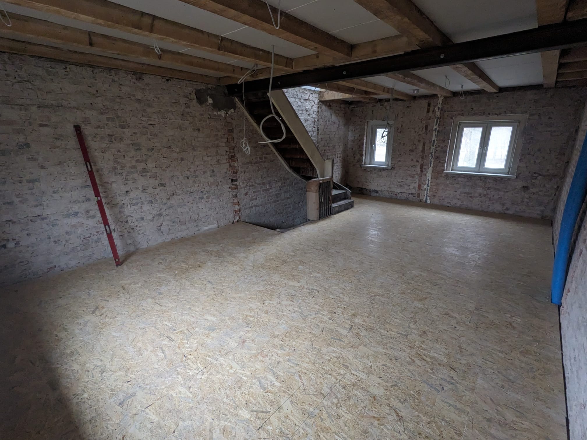Surowe wnętrze z podłogą z płyt OSB, ceglanymi ścianami i drewnianymi belkami stropowymi. Widoczny poziomica i fragment schodów. Dwa okna w głębi pomieszczenia.