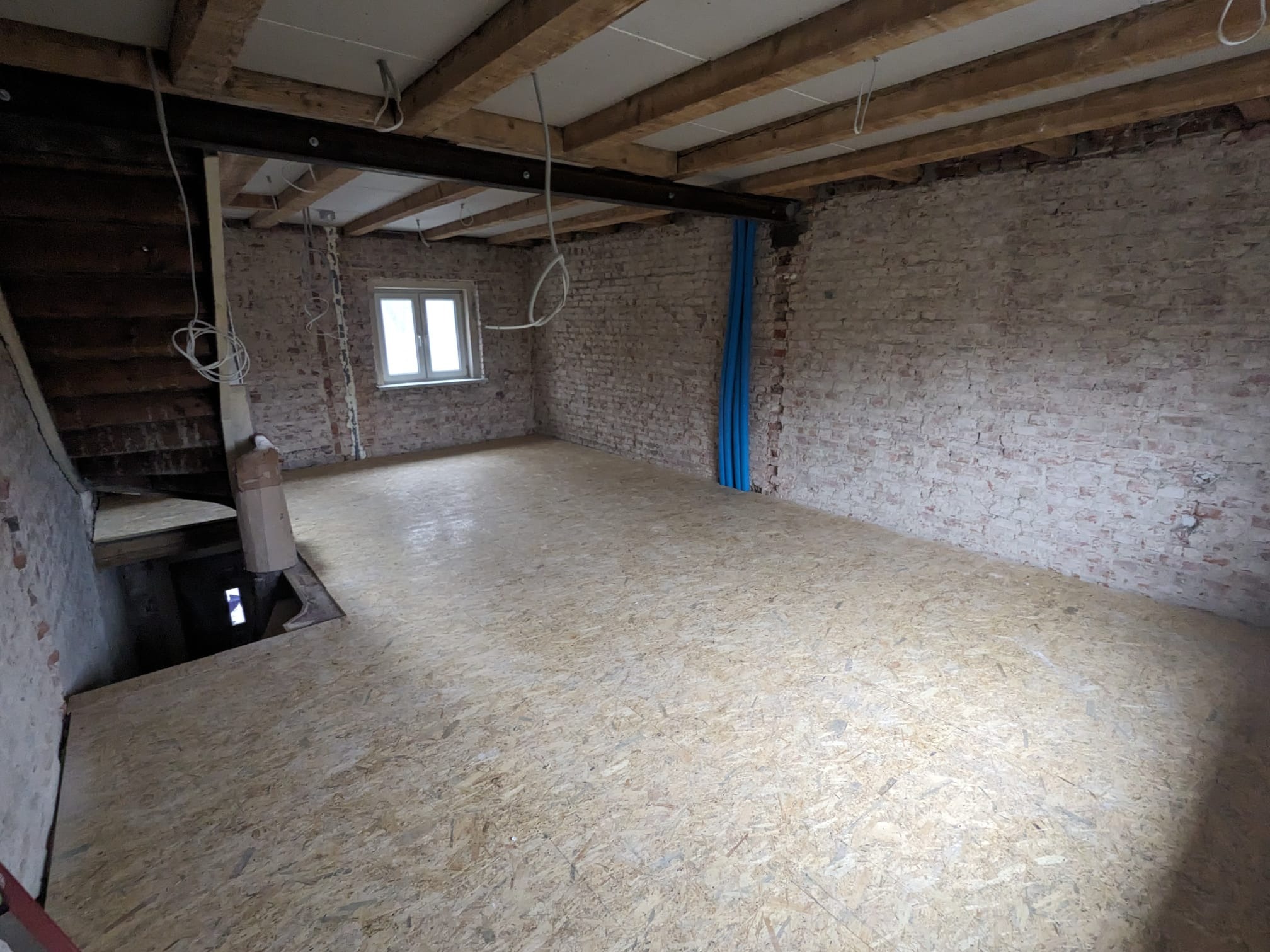 Surowe wnętrze w trakcie remontu: płyta OSB na podłodze, odsłonięta cegła na ścianach, widoczne instalacje i drewniane belki stropowe. Okno w tle.