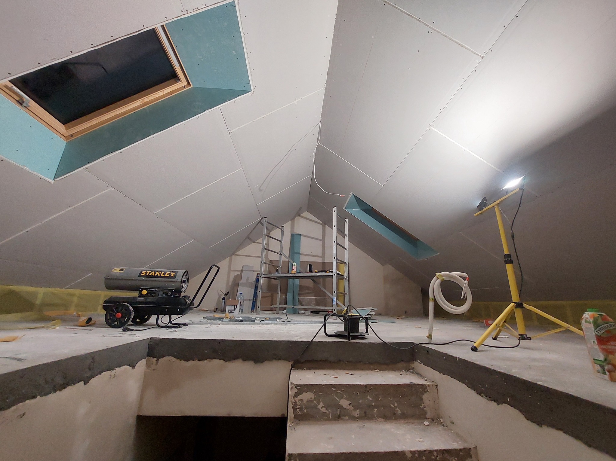 Poddasze w trakcie wykańczania: widoczne płyty karton-gips na skosach, okna dachowe, drabina i narzędzia budowlane. Oświetlenie halogenowe, surowy stan wykończenia.