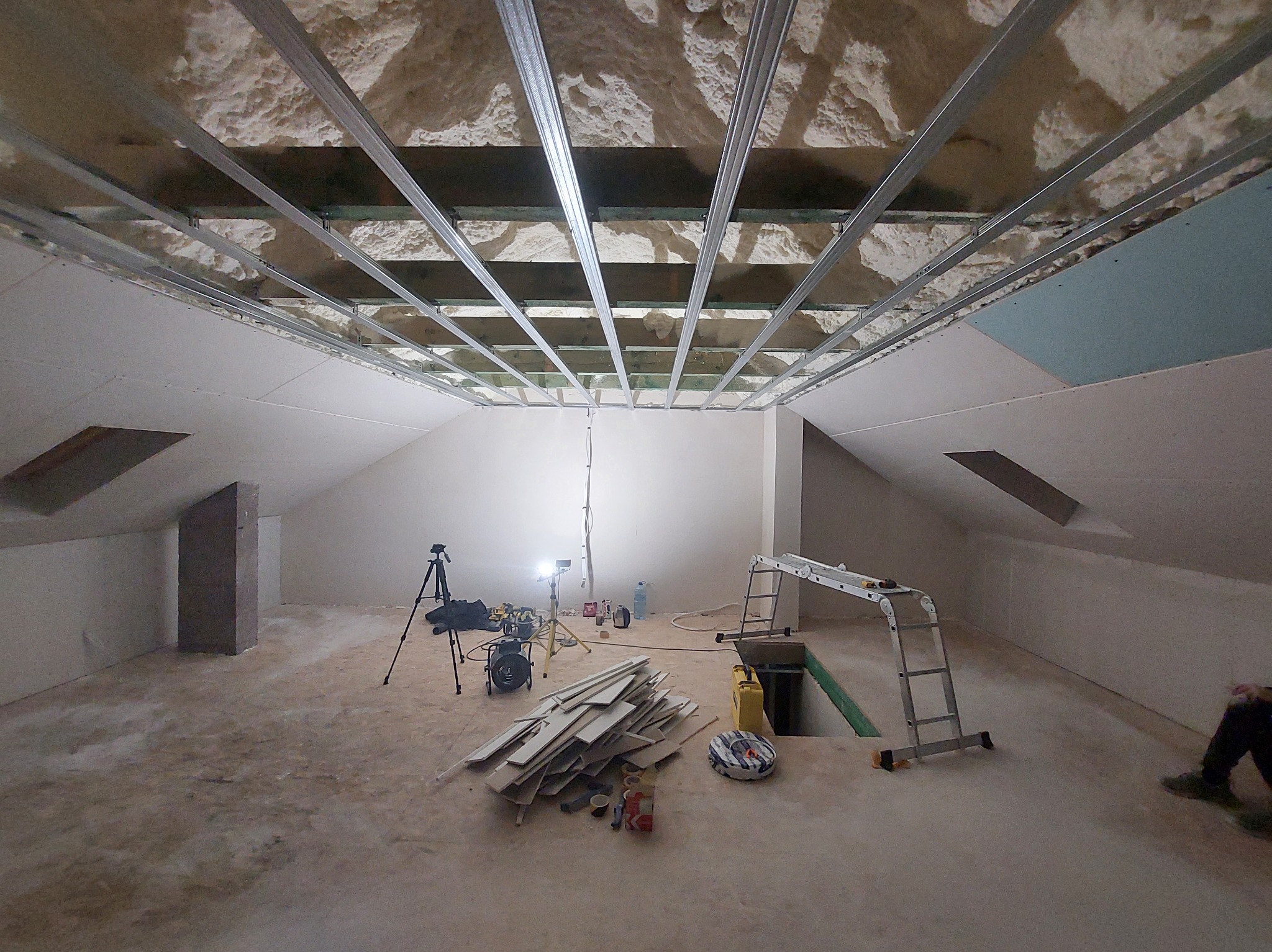Poddasze w trakcie adaptacji: widoczne metalowe profile sufitowe, izolacja z piany, częściowo zamontowane płyty gipsowe. Na podłodze narzędzia i materiały budowlane.