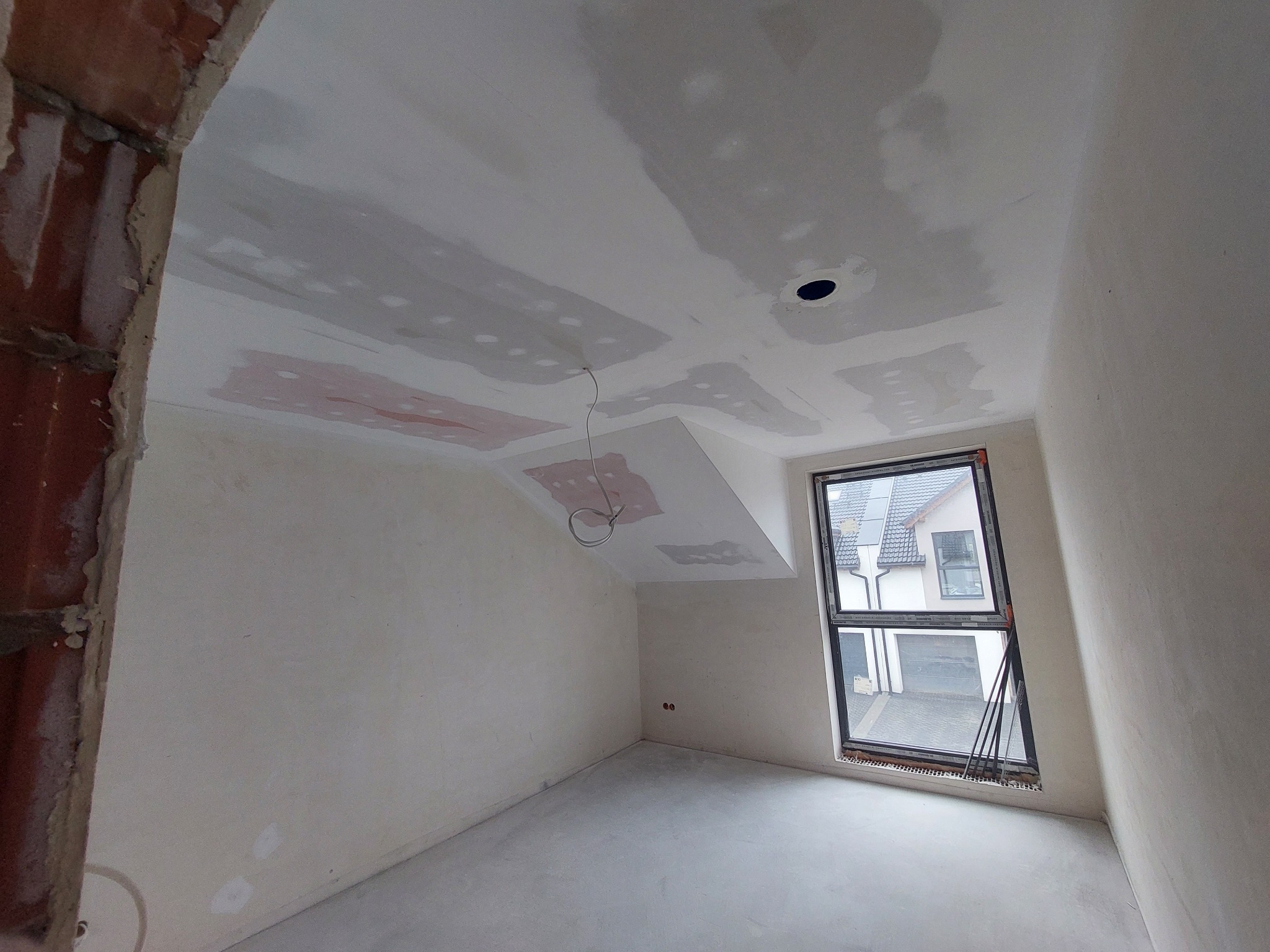 Pokój na poddaszu w trakcie szpachlowania: ściany przygotowane, widoczne łączenia płyt gipsowych. Okno z widokiem na dom. Częściowo wykończony sufit.