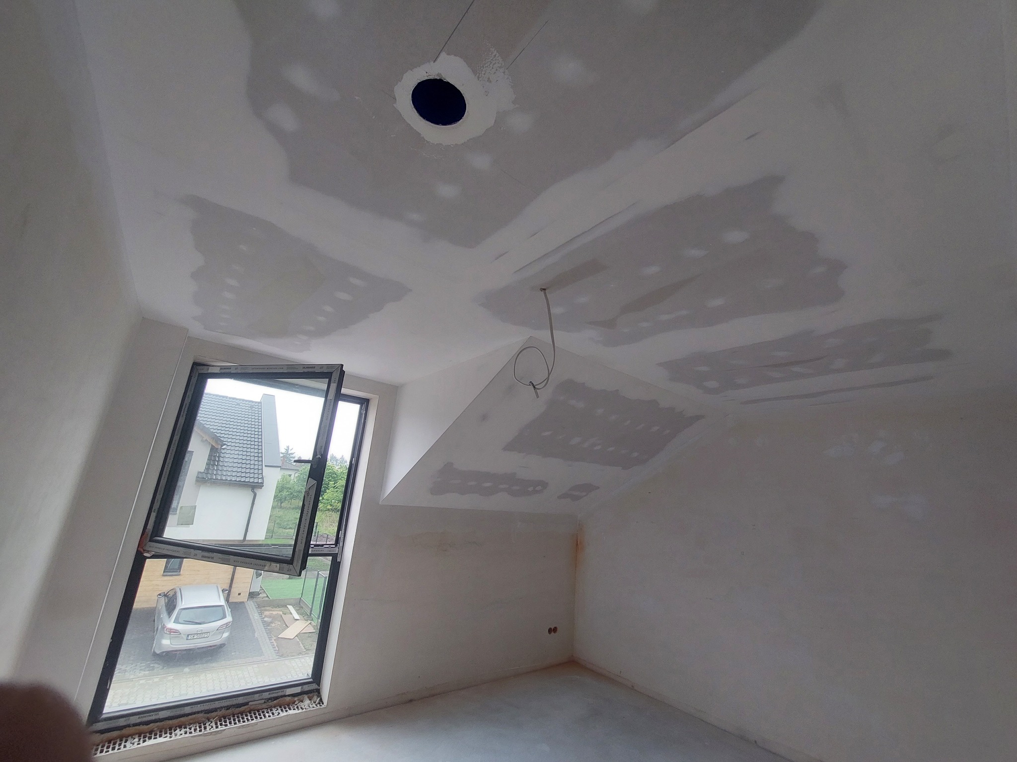 Poddasze w trakcie szpachlowania: widoczne fragmenty płyt kartonowo-gipsowych na skośnym suficie, okno dachowe z widokiem na dom i samochód. Przygotowanie do malowania.