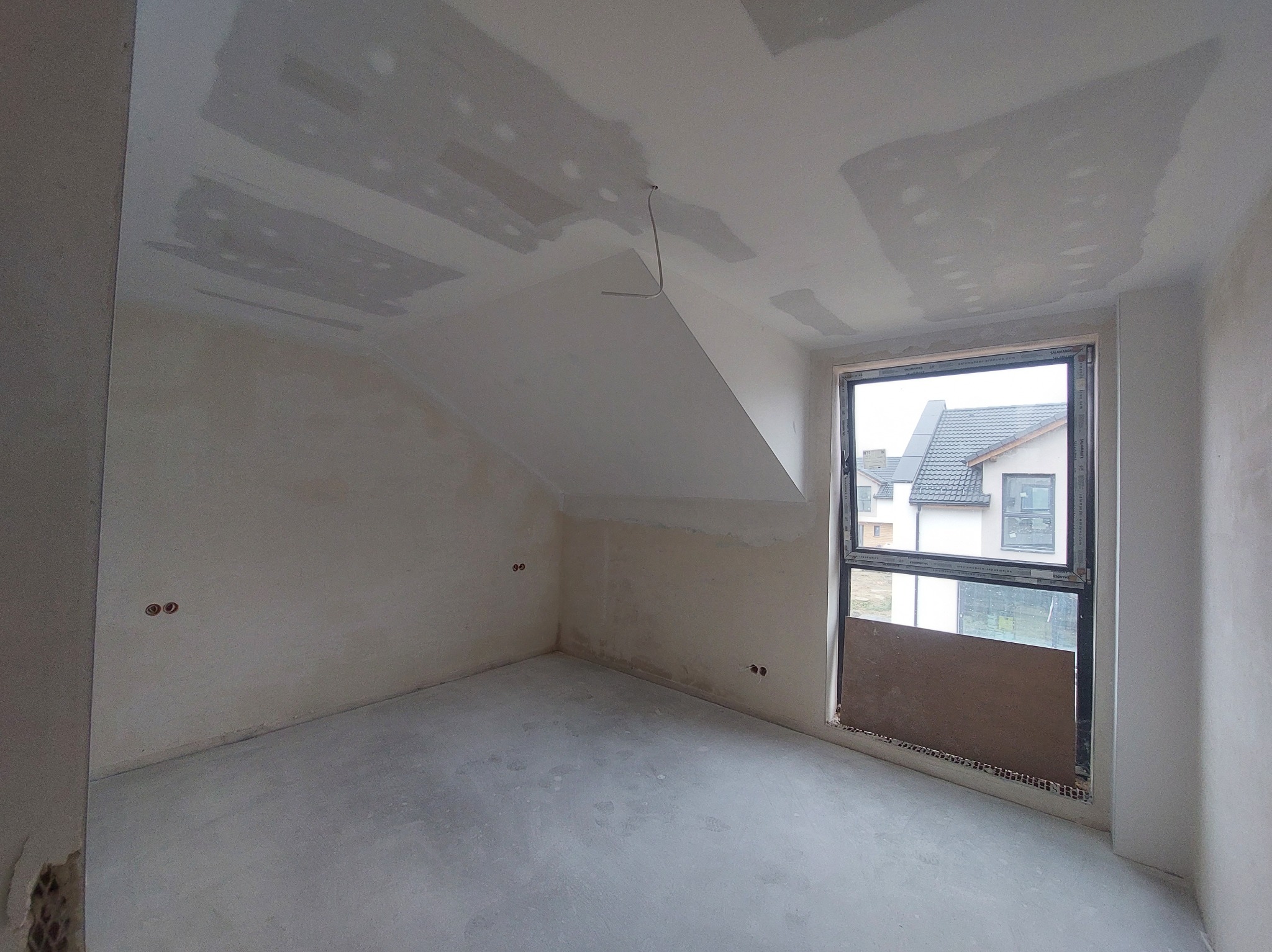 Pokój na poddaszu w trakcie szpachlowania: widoczne ślady po łatach na suficie i ścianach, surowy beton na podłodze, okno z widokiem na dom.