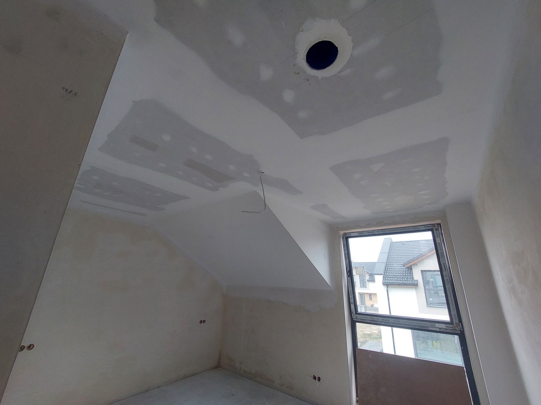 Pokój na poddaszu w trakcie prac wykończeniowych: szpachlowane ściany i sufit z otworem na oświetlenie. Widoczne okno i fragment budynku na zewnątrz.