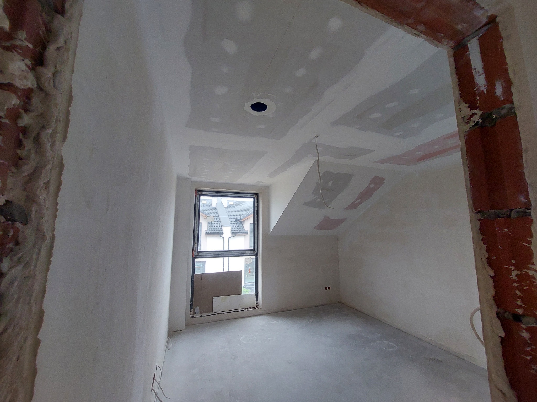 Pokój w trakcie szpachlowania: ściany i sufit pokryte gładzią, widoczne płyty karton-gips, okno i fragment surowego muru przy wejściu. W tle zabudowa jednorodzinna.