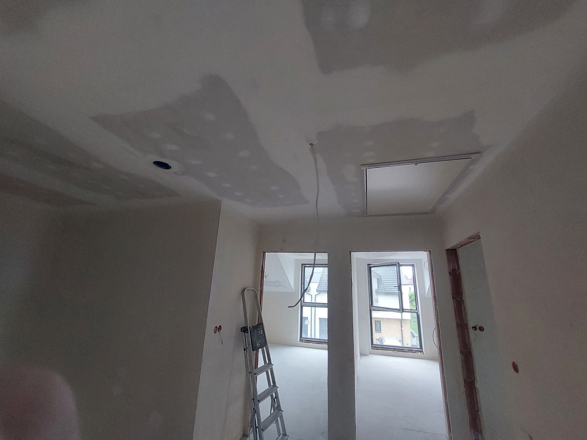 Wnętrze w trakcie prac wykończeniowych: szpachlowana sufit z widocznym włazem, ściany przygotowane do malowania, drabina, okna z widokiem na domy.