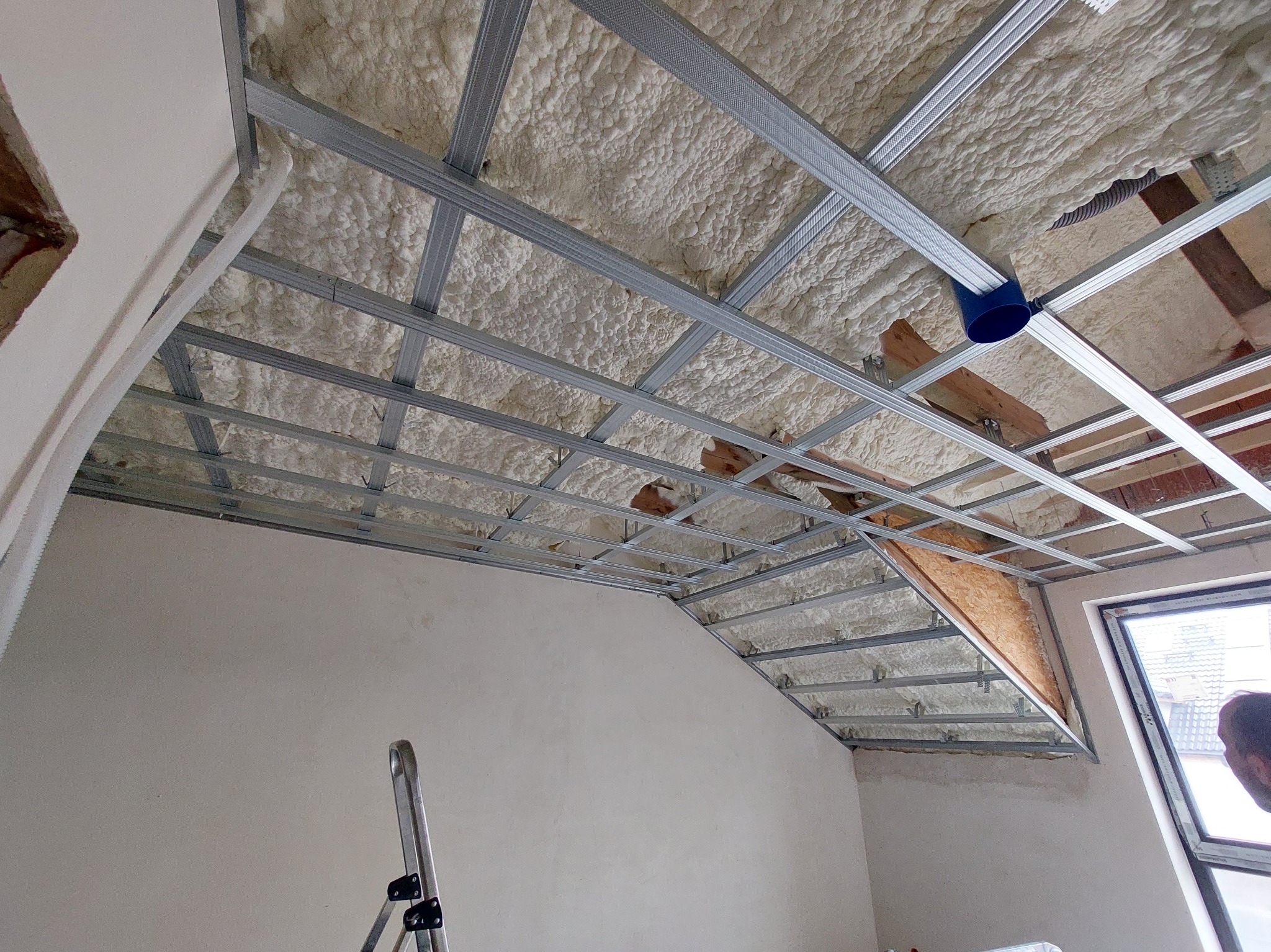 Sufit poddasza w trakcie izolacji pianą PUR i montażu metalowej konstrukcji pod płyty karton-gips, widoczna drabina i okno.