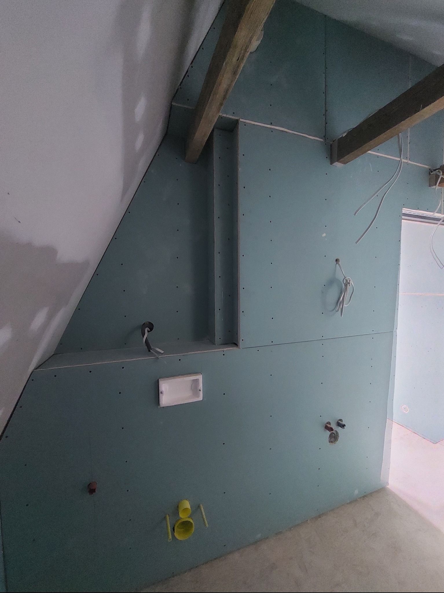 Montaż zielonej płyty gipsowo-kartonowej na poddaszu ze skosami. Widoczne drewniane belki, instalacja elektryczna i otwory na instalacje wodno-kanalizacyjne.