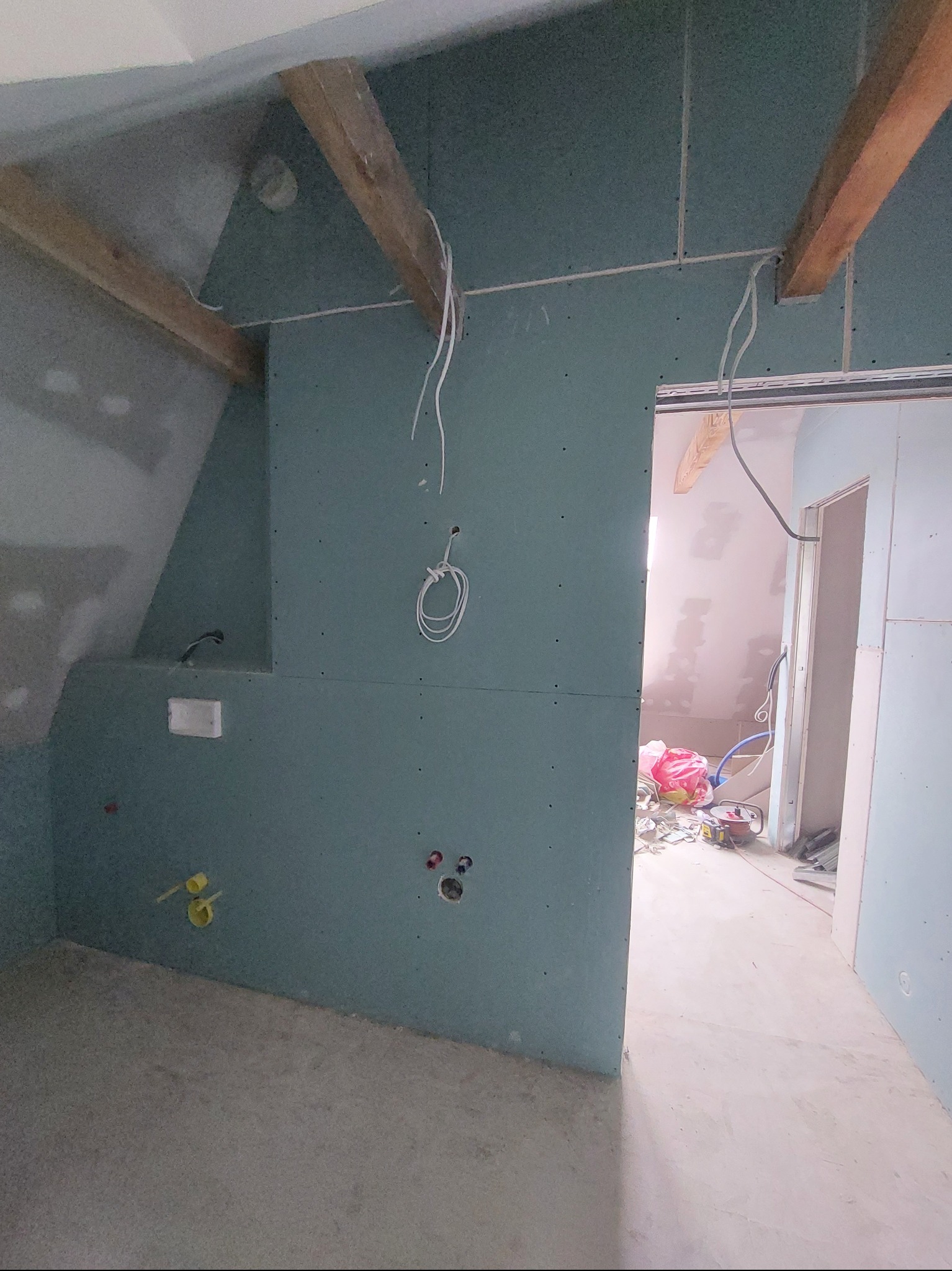 Wnętrze w trakcie prac wykończeniowych: surowe ściany z płyt gipsowo-kartonowych, widoczne instalacje elektryczne i hydrauliczne, drewniane belki stropowe.