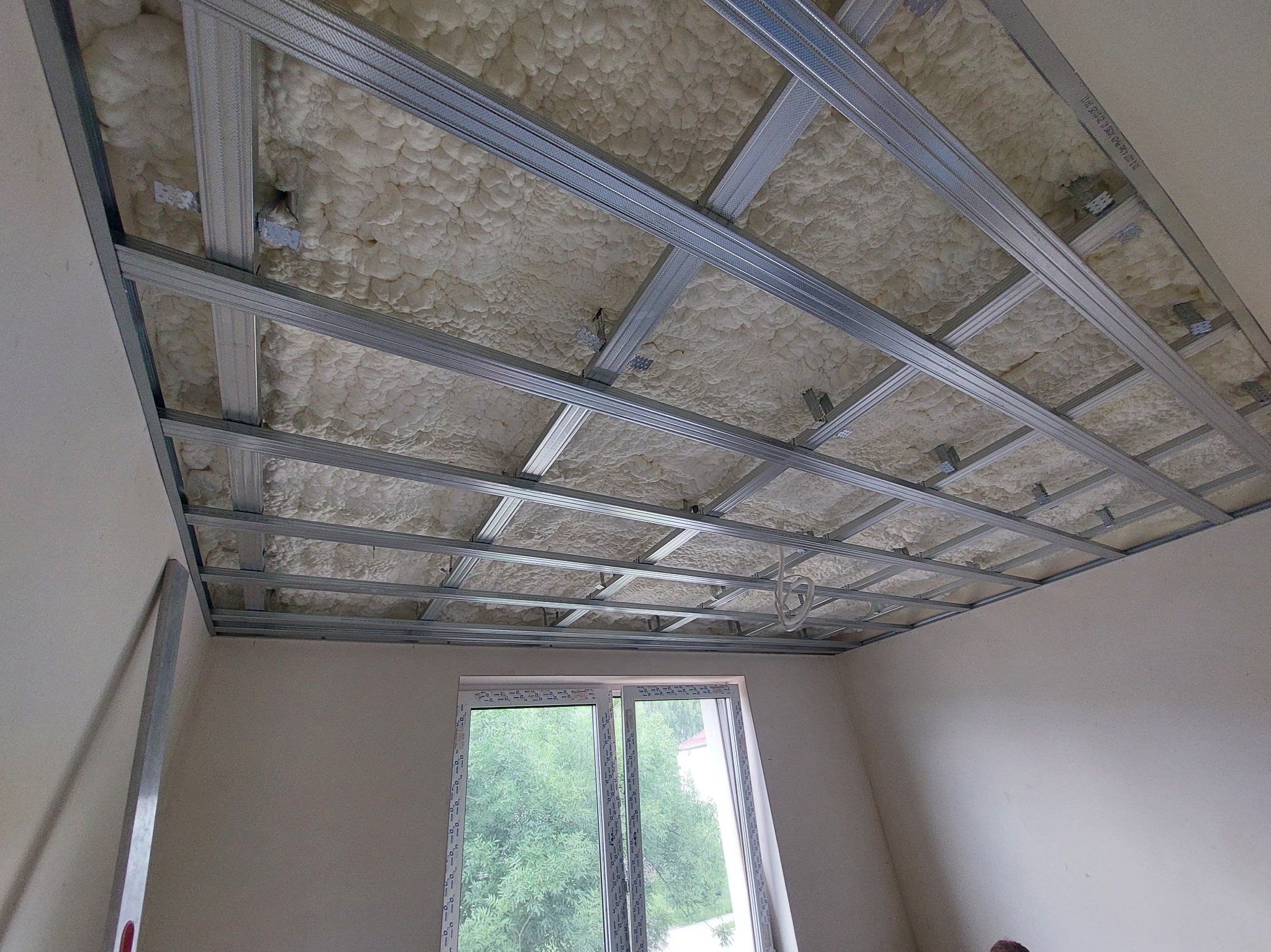 Sufit podwieszany w trakcie montażu: metalowa konstrukcja kratowa wypełniona pianką izolacyjną, widok z dołu, okno w tle. Etap prac wykończeniowych.