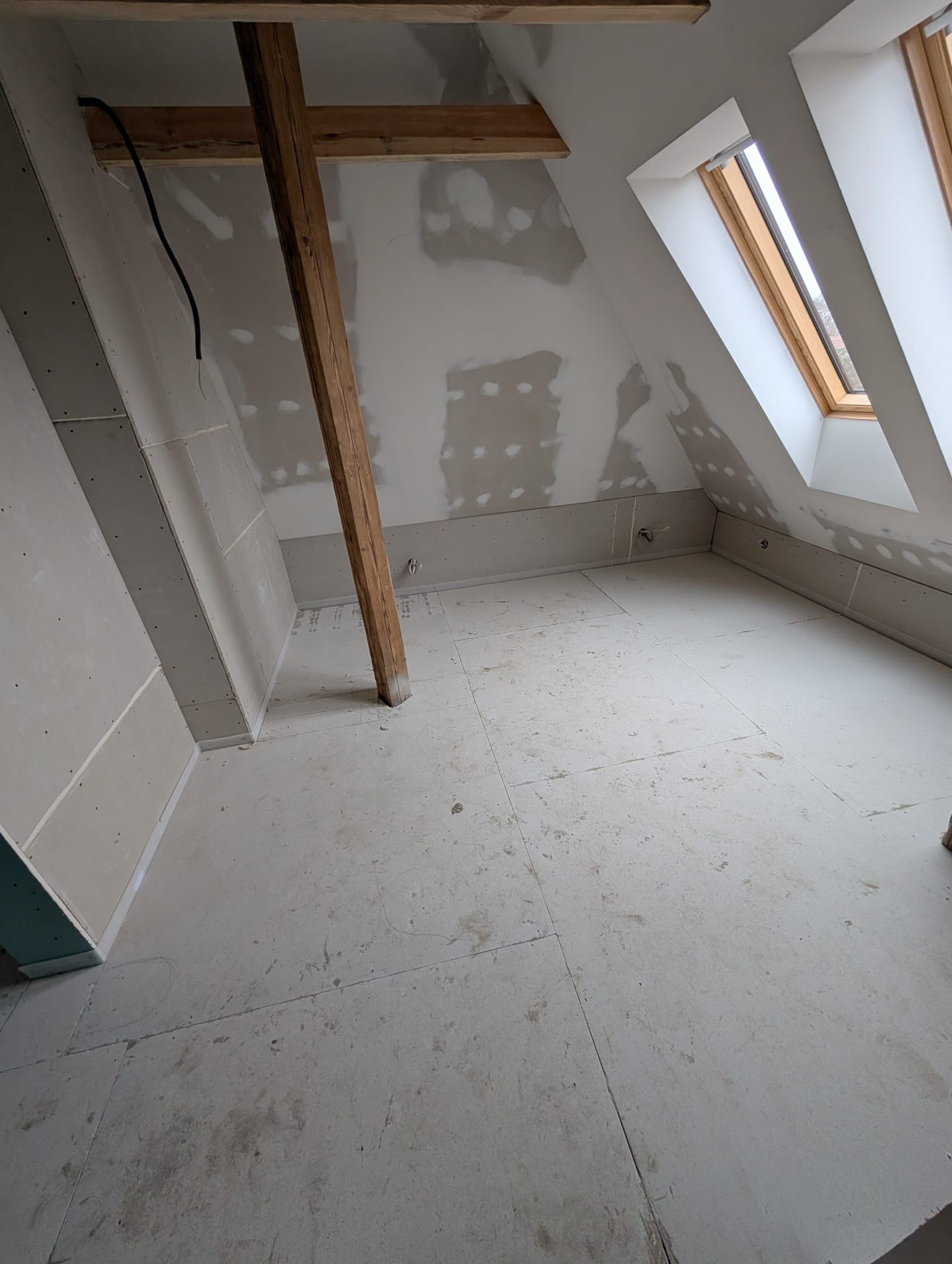 Poddasze w trakcie wykańczania: płyty karton-gips na ścianach, drewniana belka konstrukcyjna, okno dachowe. Widoczne ślady szpachlowania. Podłoga przygotowana do dalszych prac.