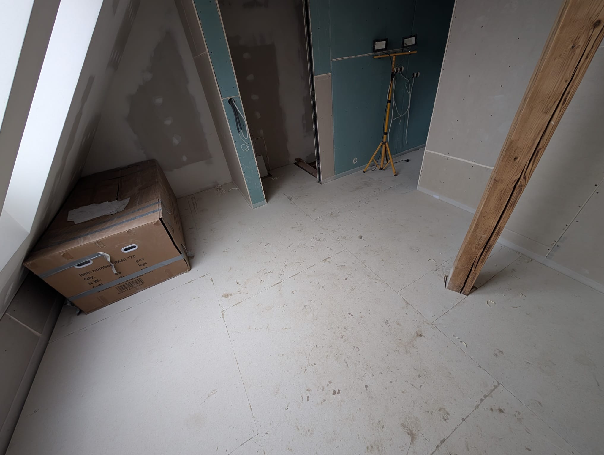 Poddasze w trakcie prac wykończeniowych: płyty gipsowe na ścianach, widoczna konstrukcja drewniana, karton na podłodze. W tle lampa budowlana na statywie.