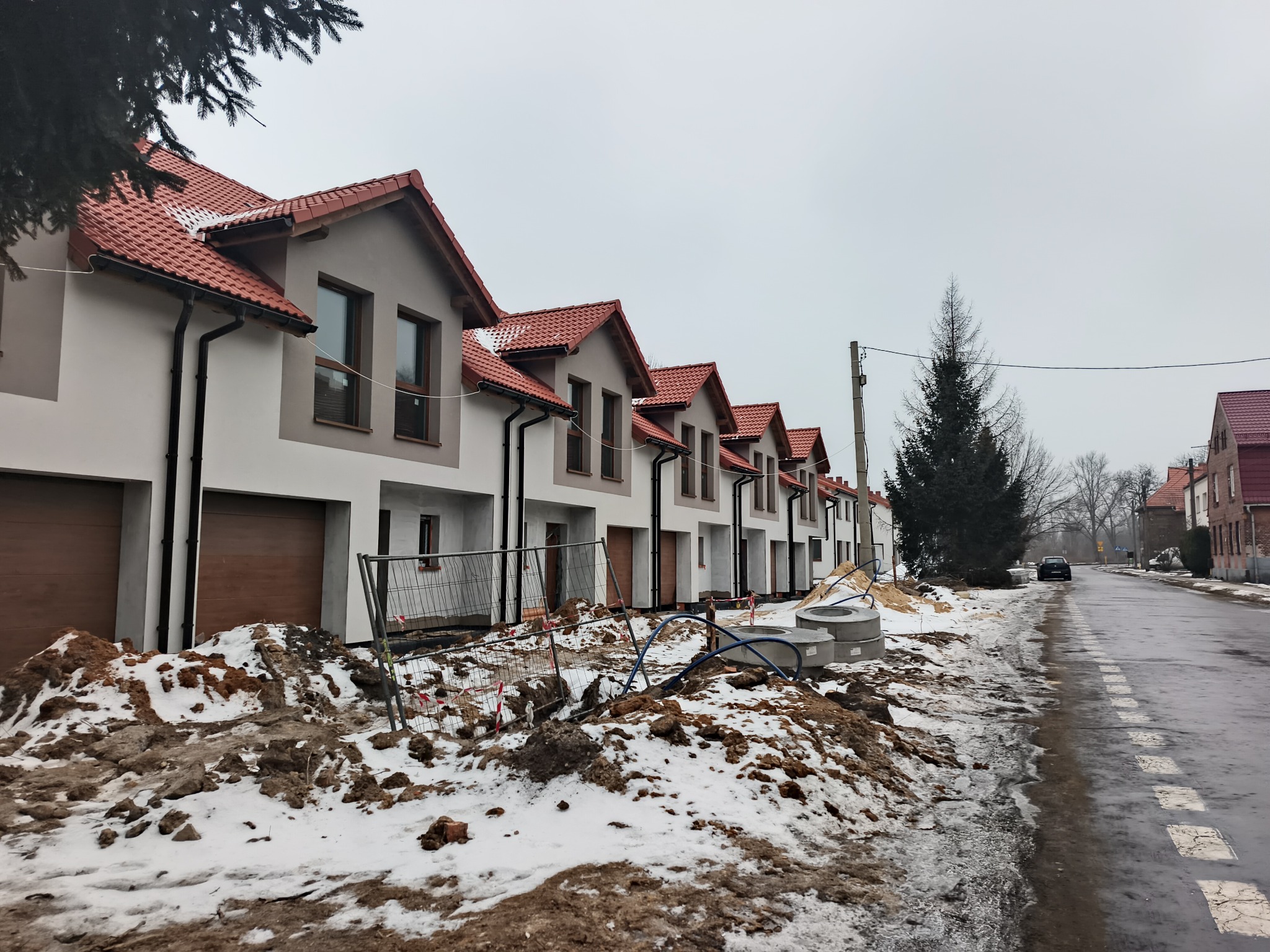 Szereg identycznych, dwukondygnacyjnych domów z czerwonymi dachami w trakcie budowy. Widoczne ogrodzenie placu budowy i zaspy śniegu. Pochmurne niebo.